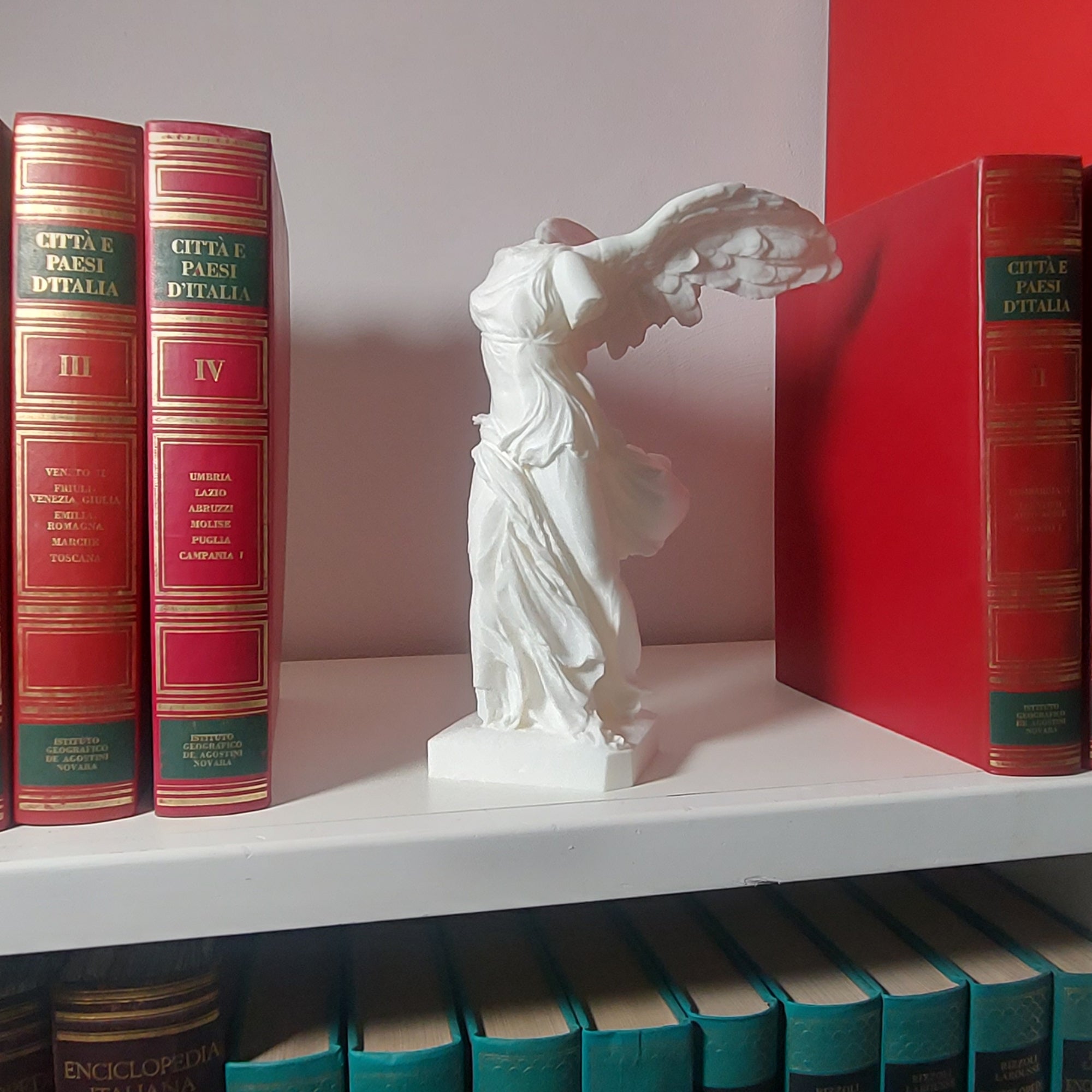 Statua della Nike in stampa 3D esposta su uno scaffale tra libri classici rossi.