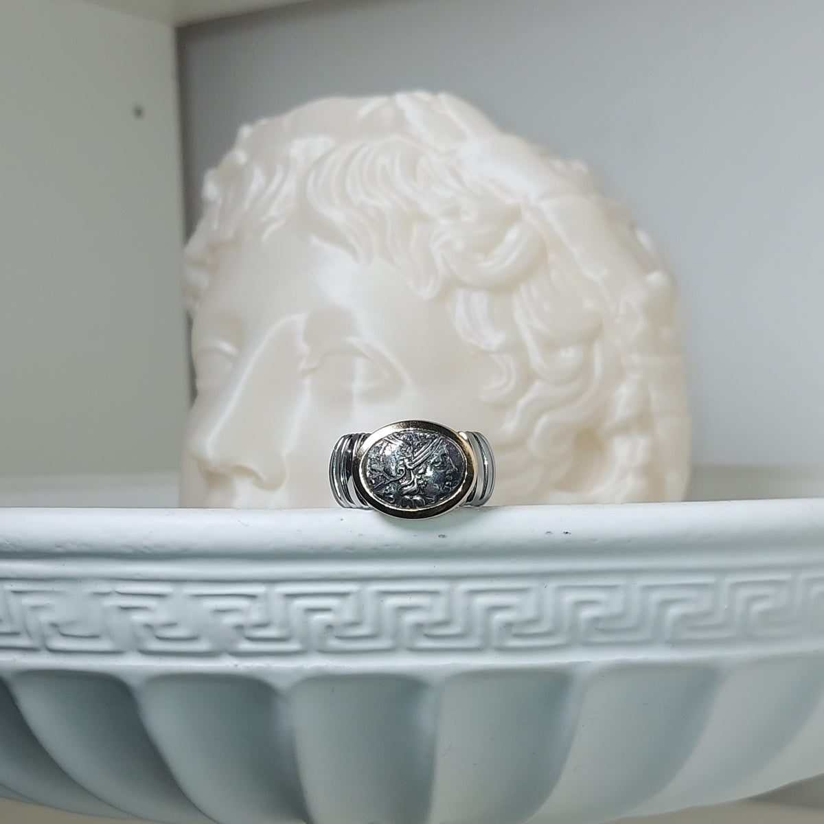 Anello in argento con riproduzione di moneta antica, esposto davanti a un rilievo di volto classico