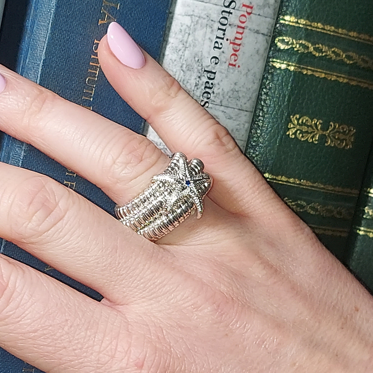 Dettaglio dell'anello a sei giri con stella marina e centro blu, indossato sulla mano davanti a libri