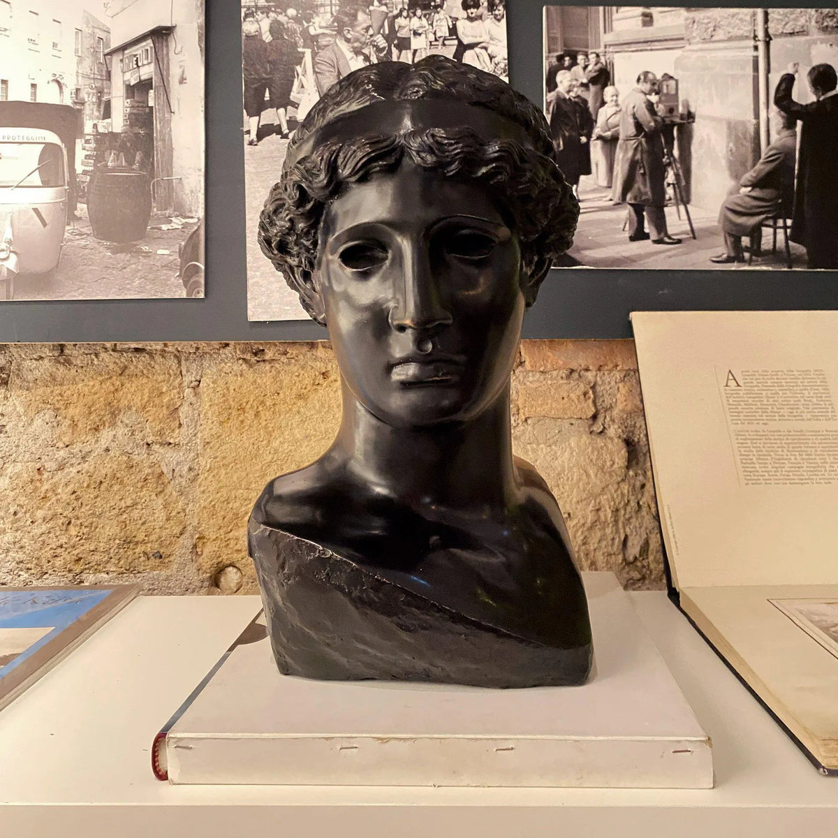 Testa di Atena Lemnia in bronzo nero esposta sopra un libro, con fotografie in bianco e nero sullo sfondo.