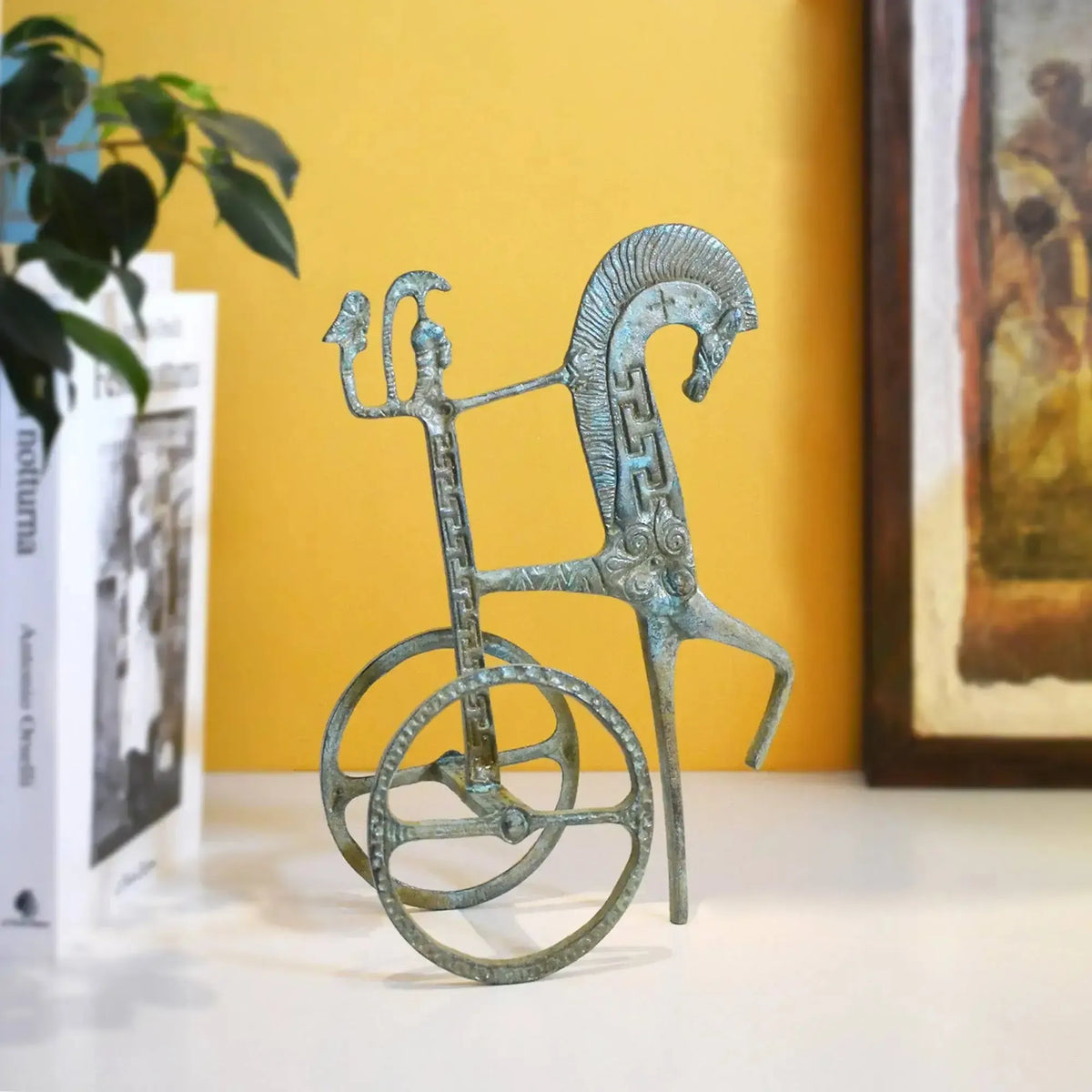 Bronzefigur einer griechischen Zweispänner-Biga mit grüner Patina, stilisiertem Pferd und dekorativen Rädern auf weißer Fläche.