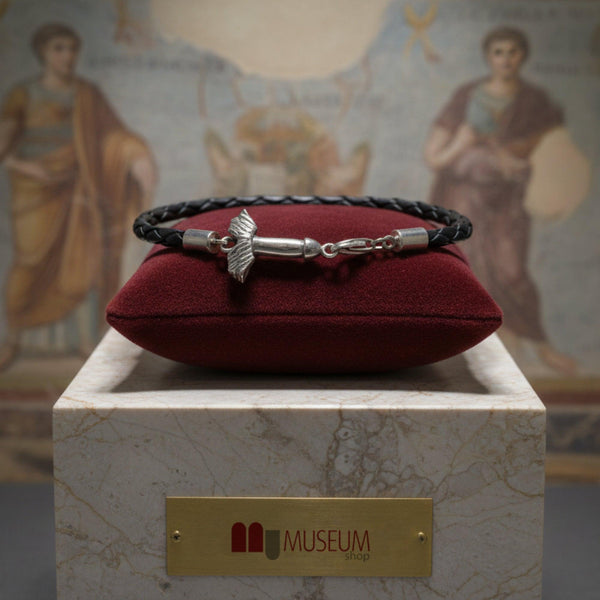 Bracciale in filo nero intrecciato con fallo alato in argento, su un cuscino rosso davanti a una parete dipinta