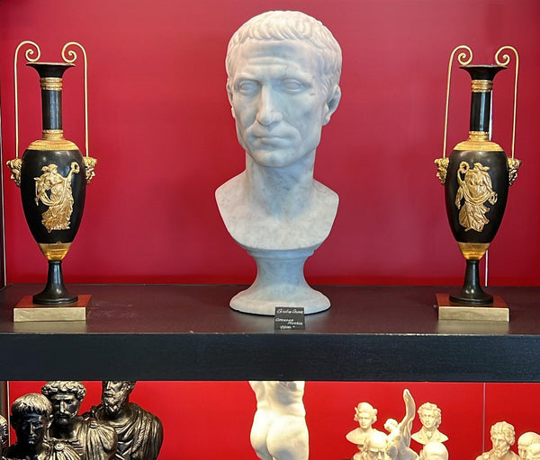 Busto di Giulio Cesare in marmo bianco di Carrara,  vista frontale su un piano di legno  vicino a vasi dorati.