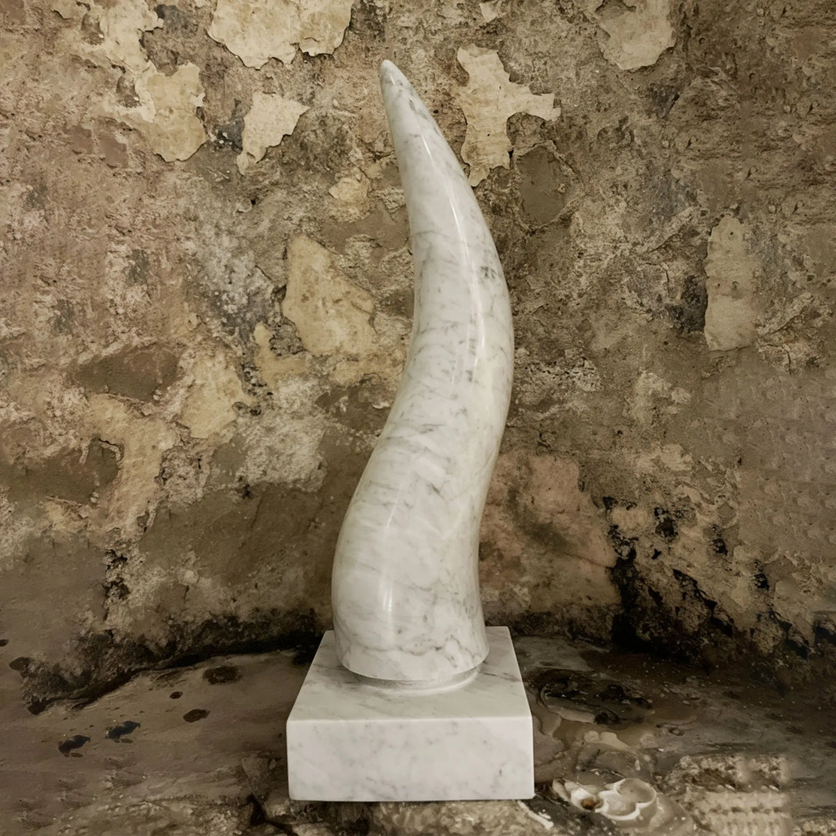Corno portafortuna in marmo di Carrara su base quadrata liscia, davanti a una parete rustica.