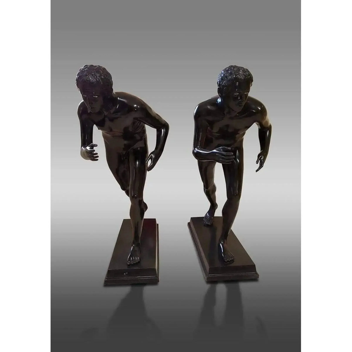 Coppia di statue del corridore di Ercolano in bronzo scuro, raffigurate in posa dinamica su basi rettangolari, viste frontalmente su fondo grigio.