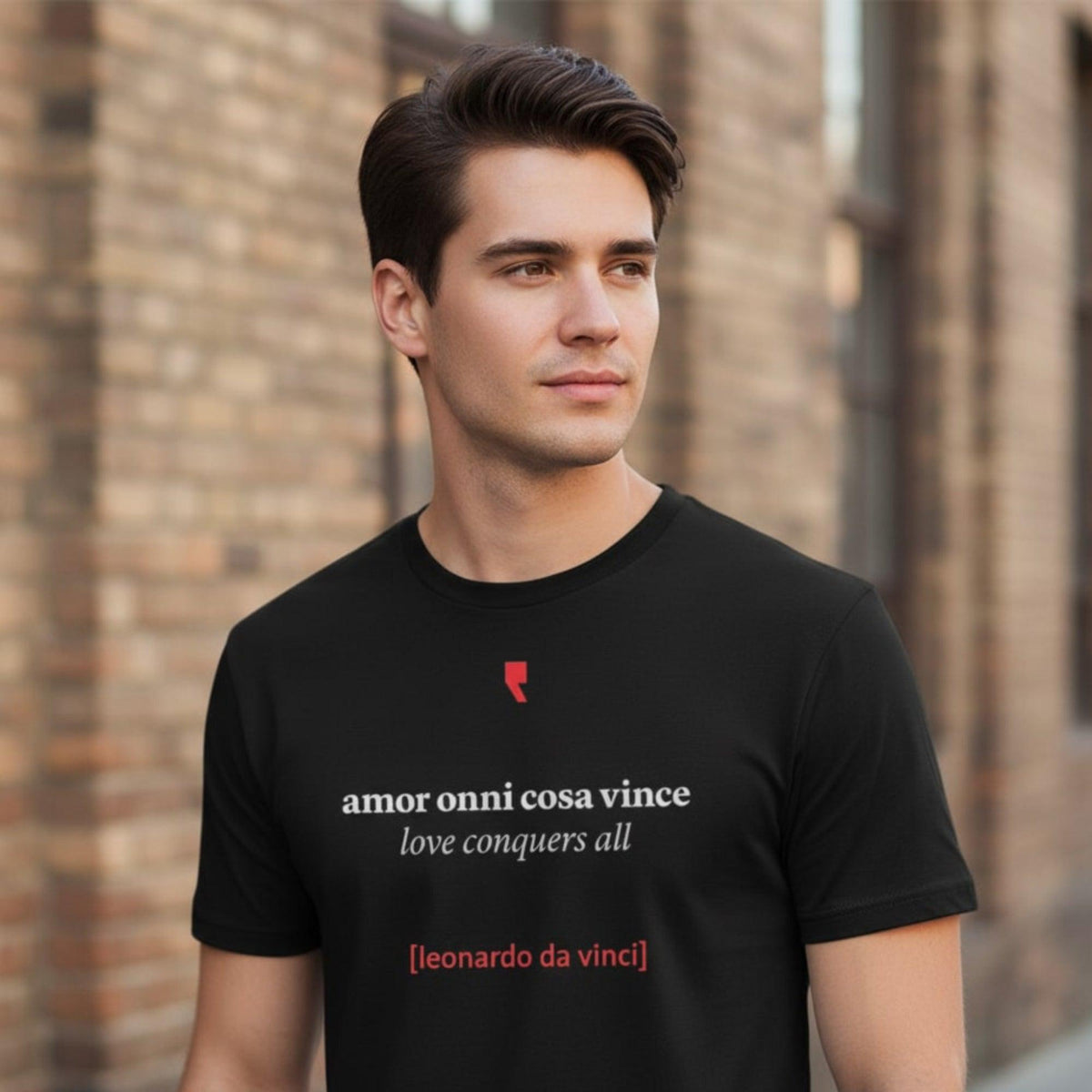 Uomo con maglietta nera “Amor onni cosa vince”, davanti a un muro in mattoni