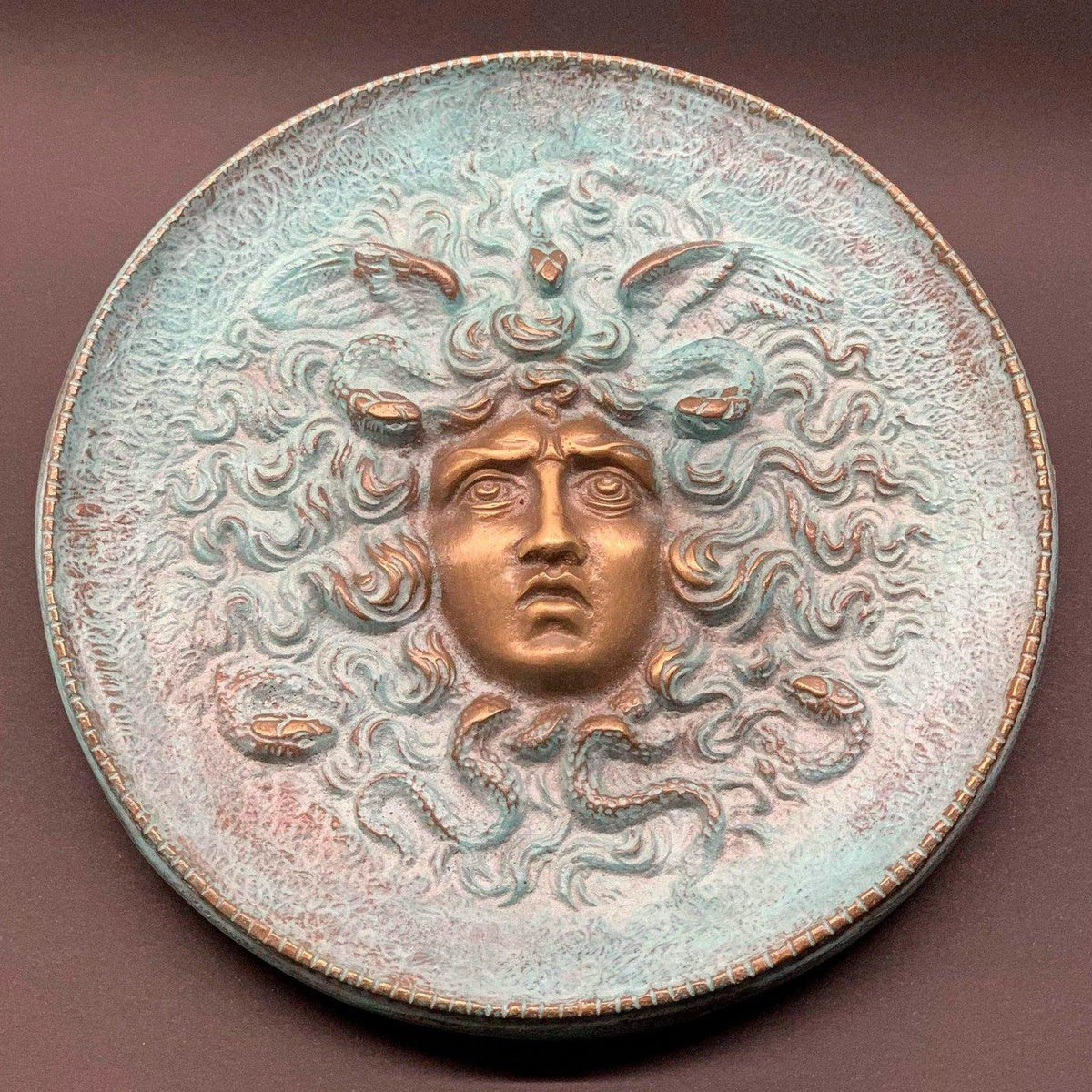 Piatto in bronzo con volto di Medusa in rilievo, serpenti e patina verde, su fondo marrone.