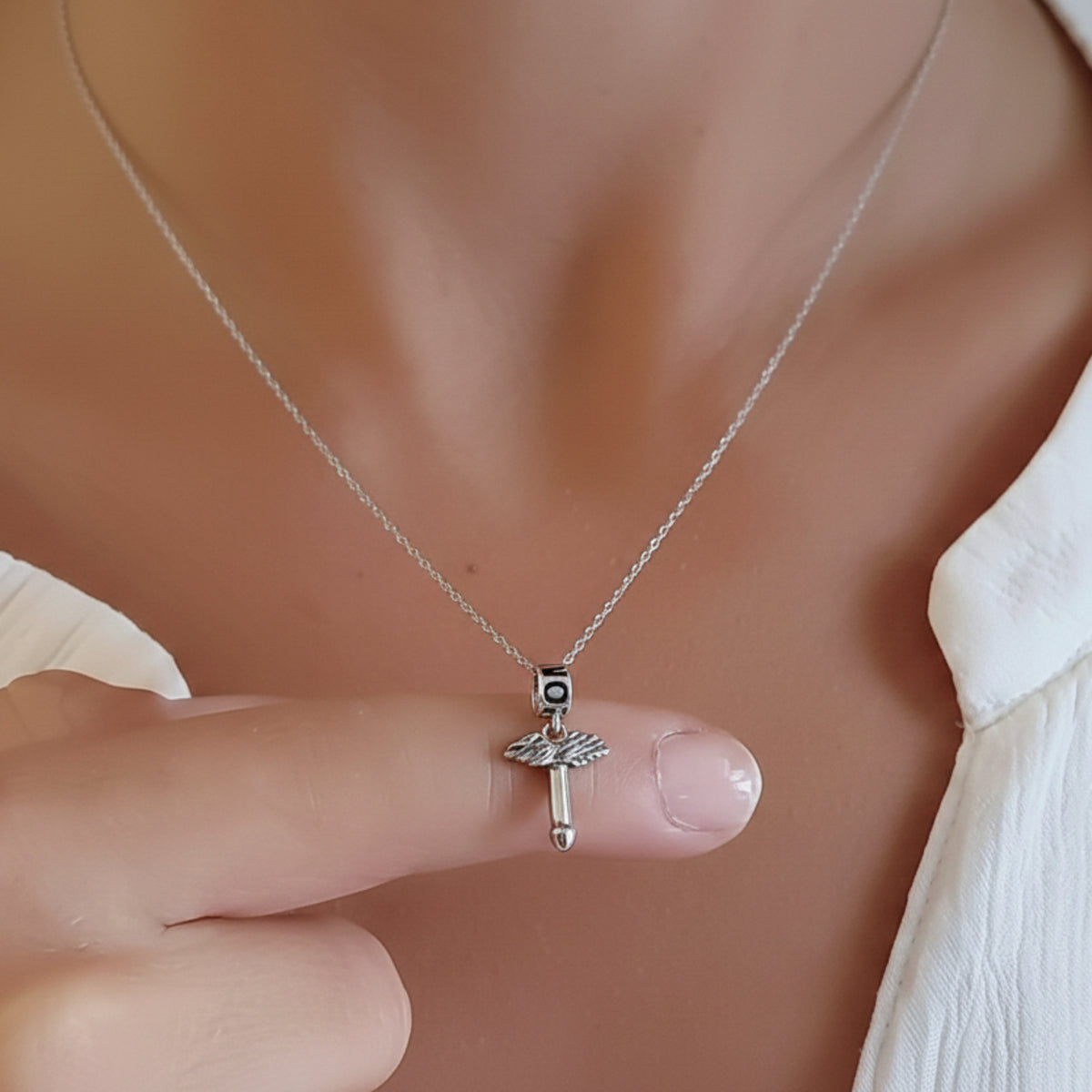 Pendente con fallo alato in argento, indossato con una catenina e appoggiato su un dito