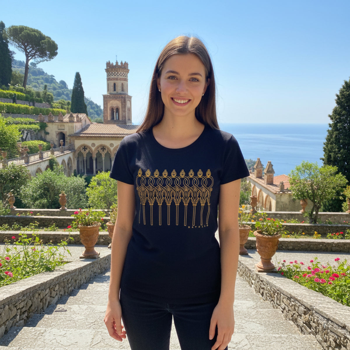 T-shirt nera da donna con archi intrecciati dorati, indossata in un giardino con vista sul mare.