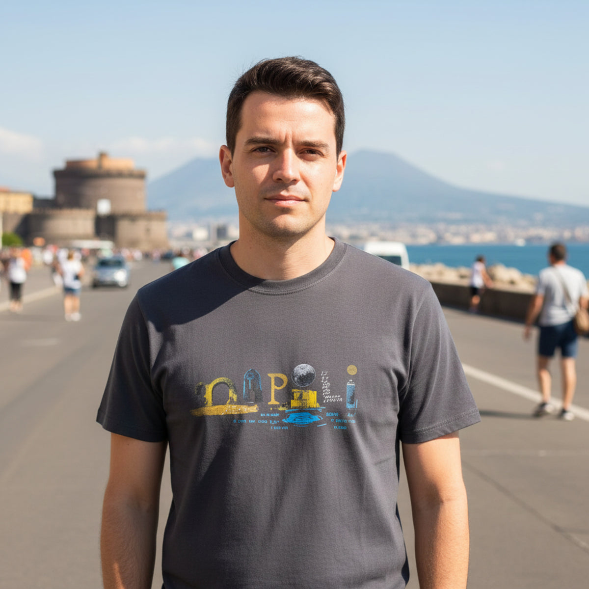 T-shirt grigia da uomo con scritta multicolore «Napoli», indossata sul lungomare con mare e montagna sullo sfondo.