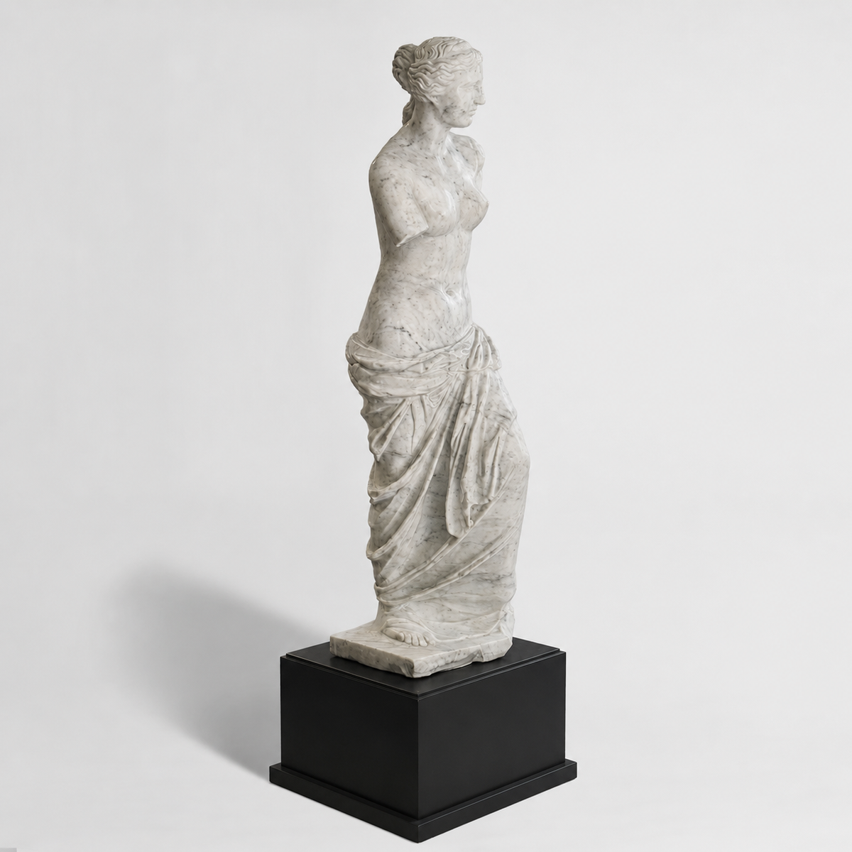 Venere di Milo in marmo di Carrara, vista di profilo su piedistallo nero e fondo grigio chiaro.