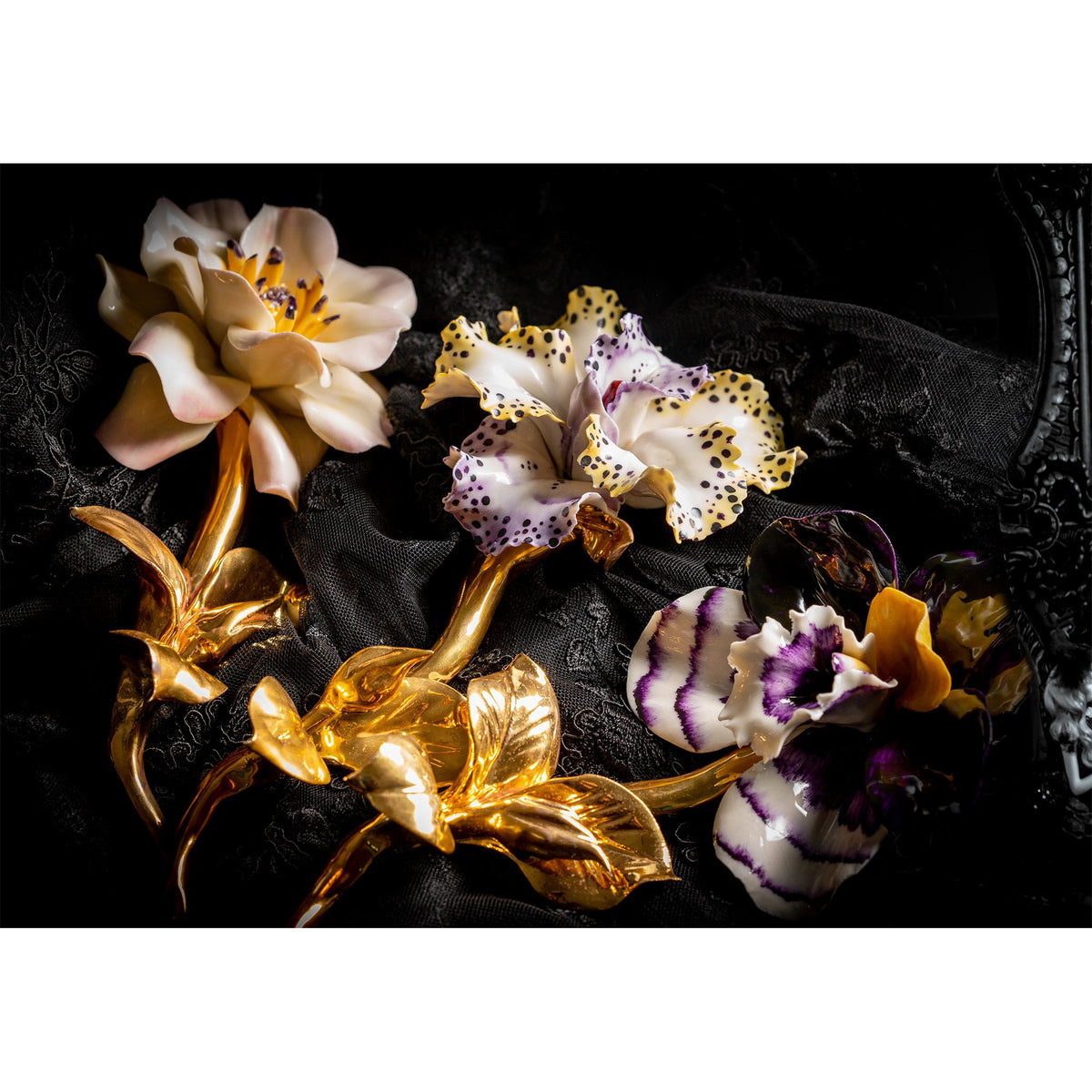 Tre fiori decorativi con gambi dorati su tessuto nero, con camelia dai petali chiari a sinistra.