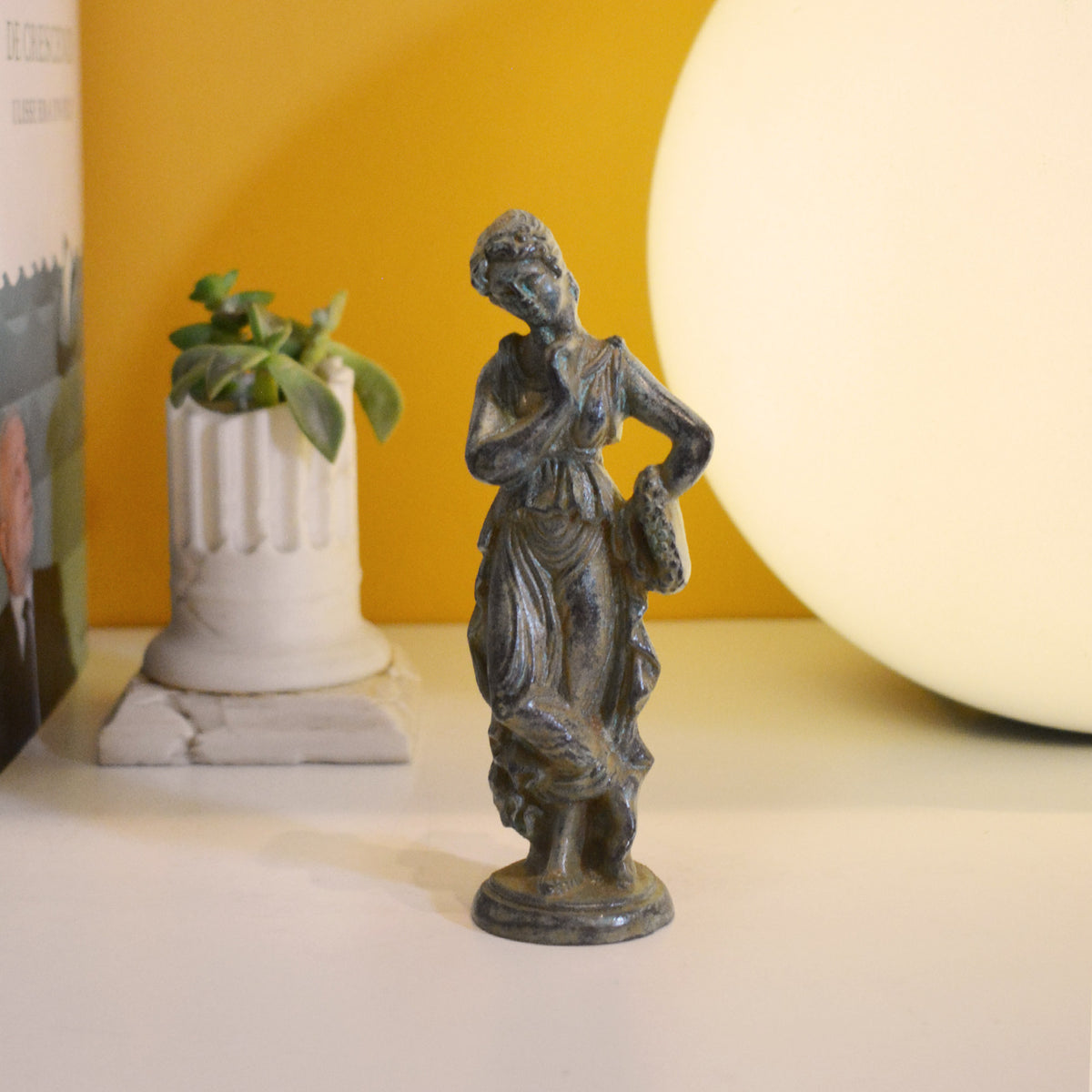 Römische Frau aus Bronze mit grüner Patina, drapiertem Kleid und dekorativem Sockel, fotografiert in einer Umgebung mit Pflanze und warmem Licht im Hintergrund.
