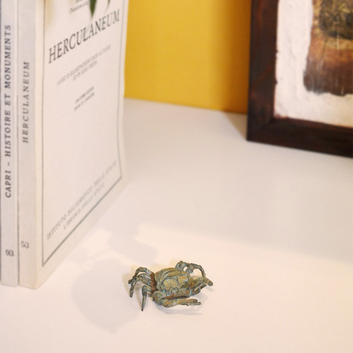 Bronzekrabbe mit grüner Patina, dekorative Figur mit Scheren und Beinen, fotografiert in einer Umgebung mit Büchern und gelber Wand im Hintergrund.
