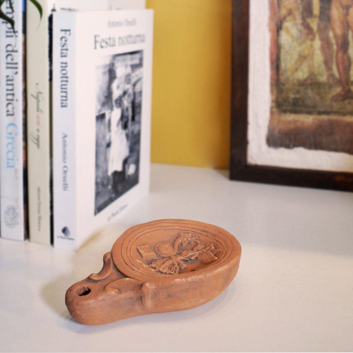 Lucerna Dea Fortuna in terracotta su un ripiano bianco accanto a libri e un quadro