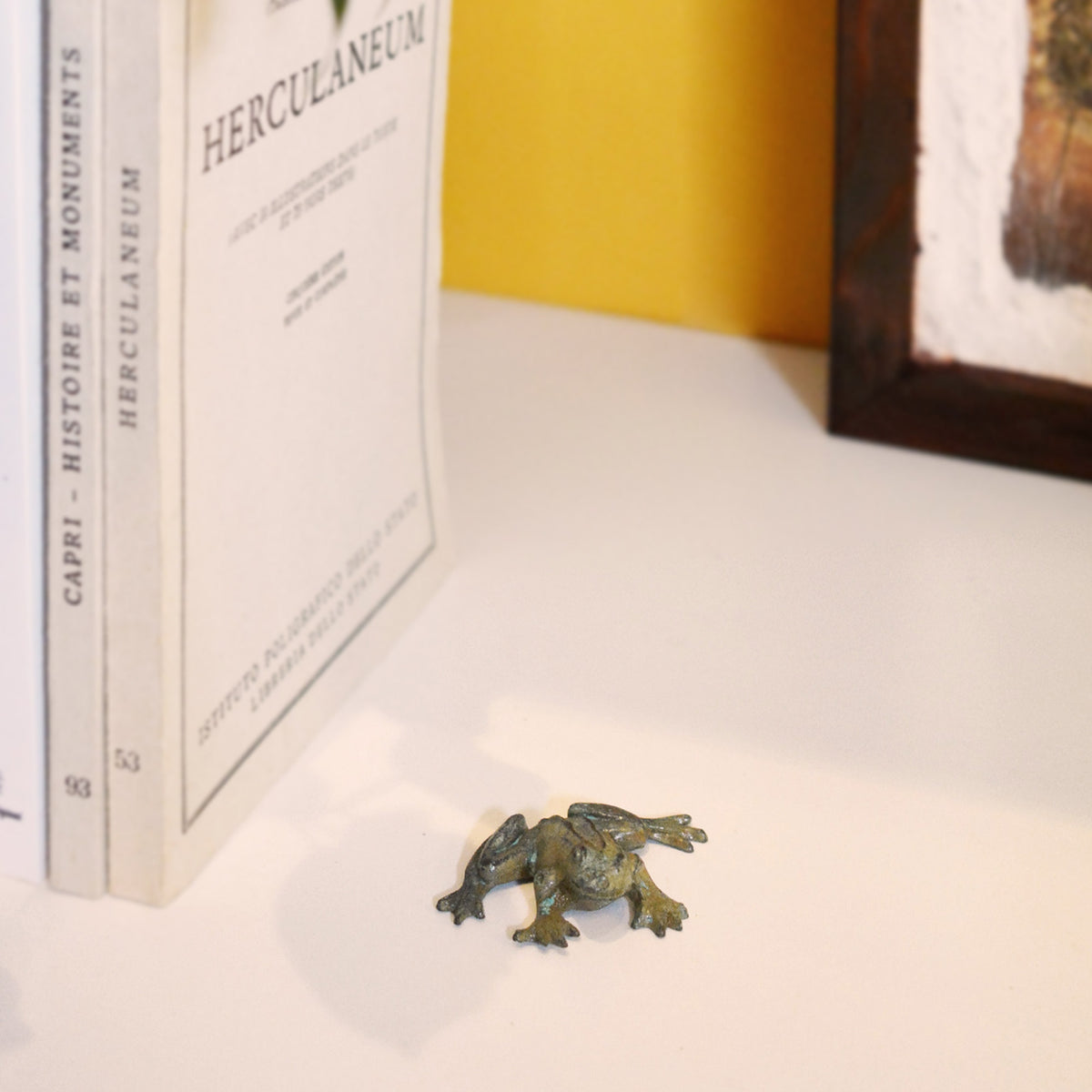 Escultura decorativa de rana de bronce con pátina verde-azulada, fotografiada sobre una superficie blanca en un ambiente con libros al fondo.