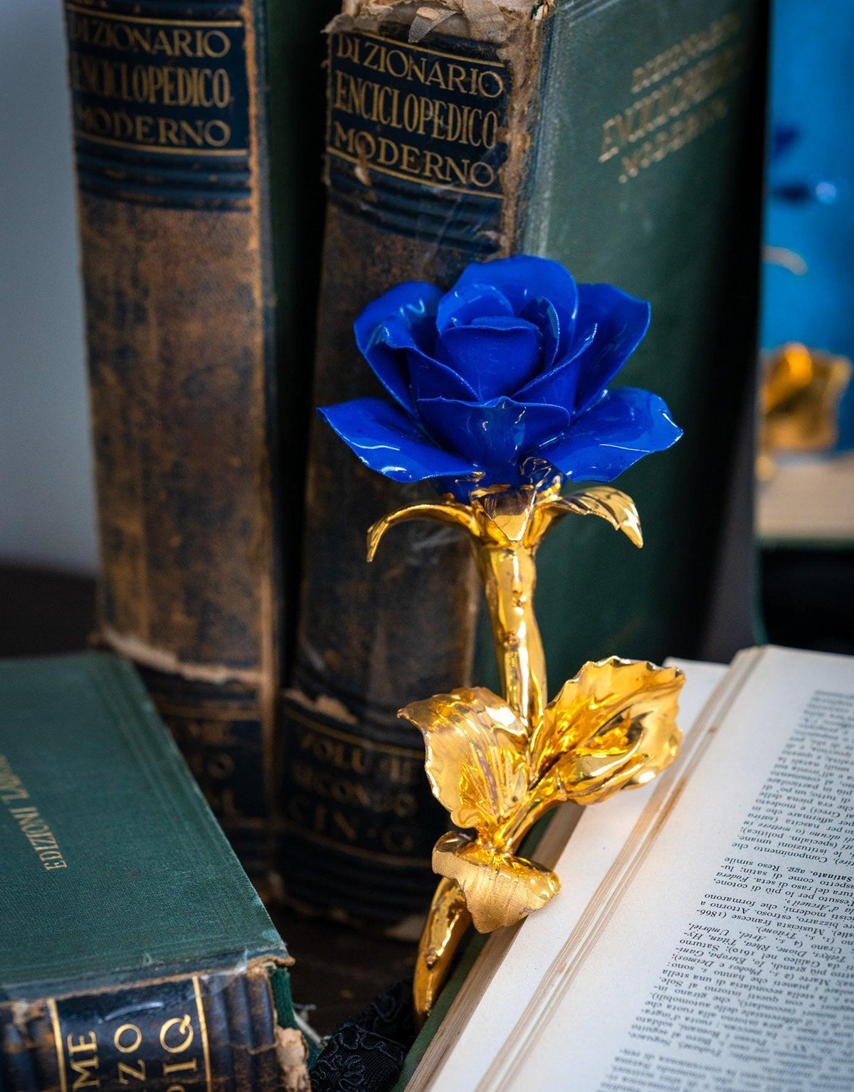 Rosa blu in porcellana di Capodimonte con gambo dorato, appoggiata su un libro aperto accanto ad altri volumi.