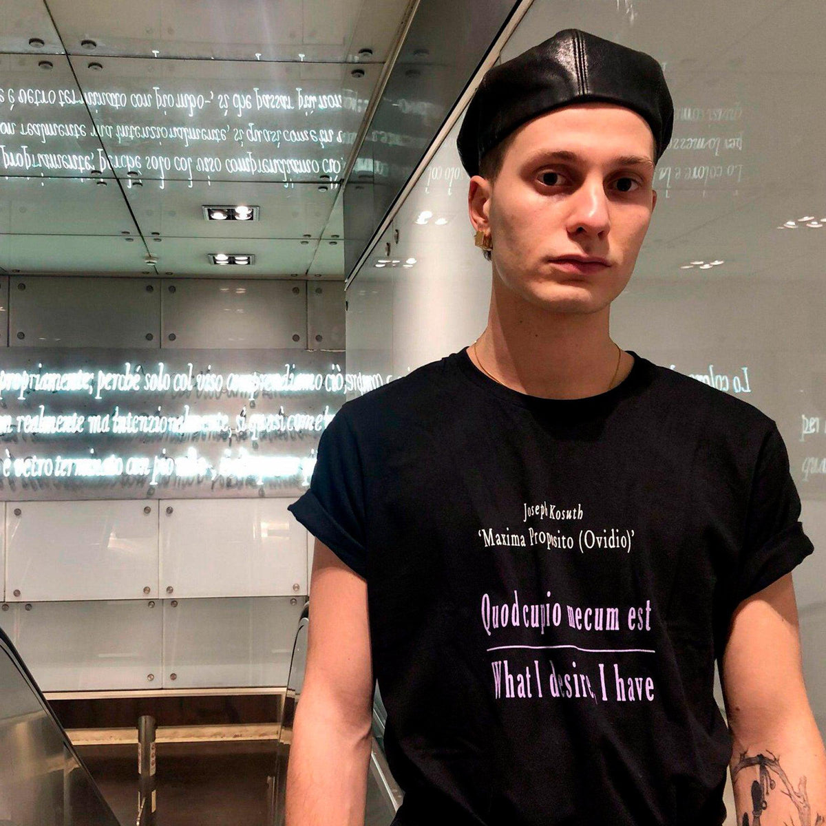 T-shirt nera da uomo con scritta viola «Quod cupio mecum est», indossata in un interno con scritte sulle pareti.